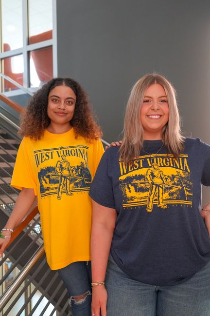 Two female students in stairwell with 2024 official wvu fan shirt in gold and blue