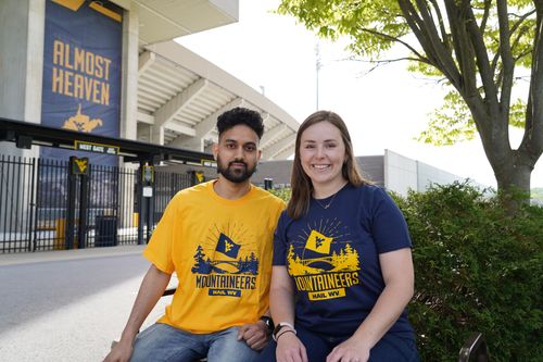 Male and female sitting on bench in front of WVU football stadium wearing a gold and navy WVU Fan shirt
