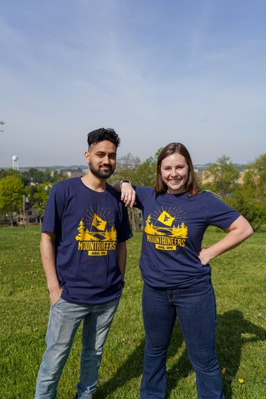 Male and female in navy wvu shirts with grass and blue skies in background